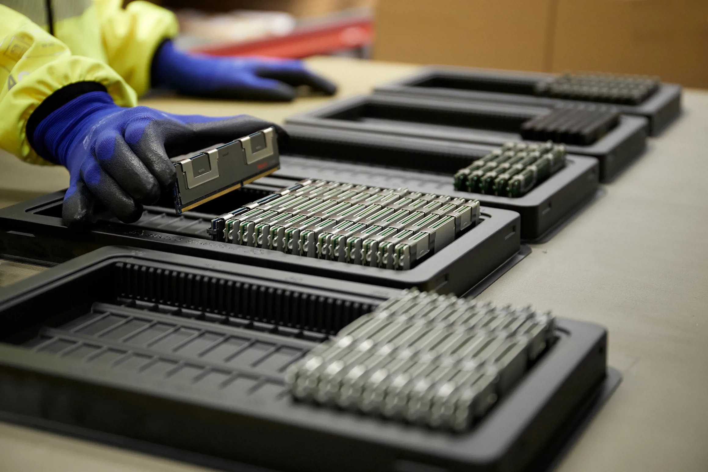 En person med blå handskar hanterar datorminnesmoduler som ligger sorterade i svarta plastbrickor på ett bord.