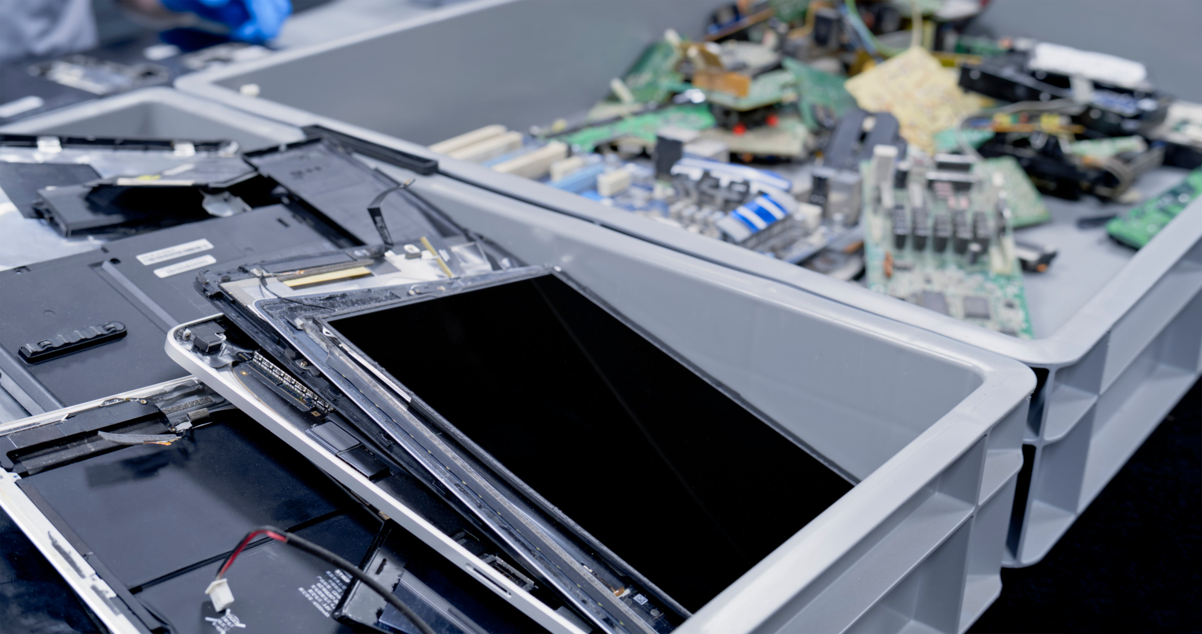 Isärtagna elektroniska enheter i grå plastlådor, inklusive surfplattor och kretskort, i en återvinnings- eller reparationsanläggning.